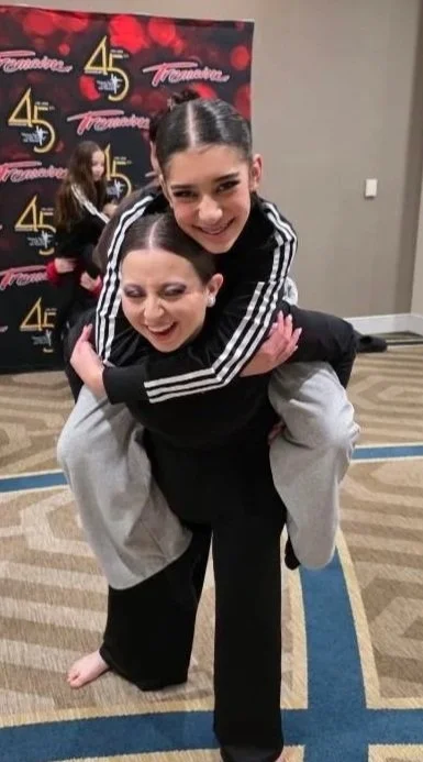 Two smiling girls, one giving the other a piggyback ride at an indoor event with a black and gold backdrop in the background.
