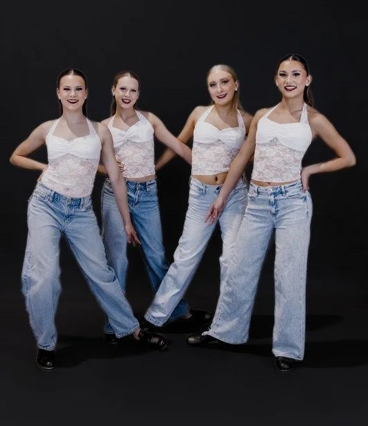 Four young women in matching white lace tank tops and high-waisted jeans pose together against a black background.