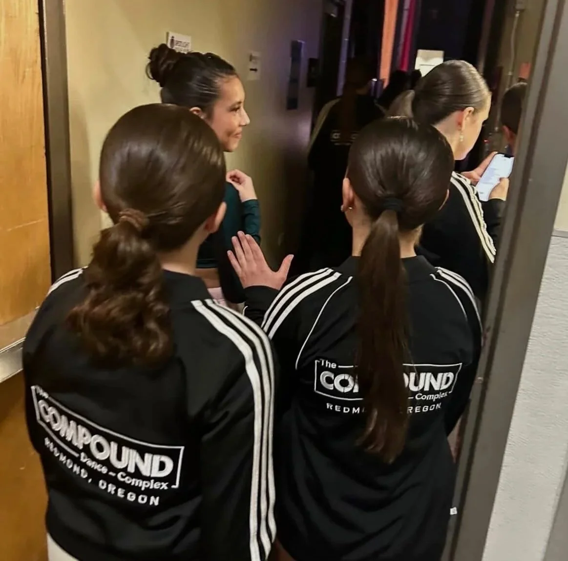 Group of women in black jackets with white stripes and 'The Comground' logo on back, standing in line in a hallway, with one woman using her phone.