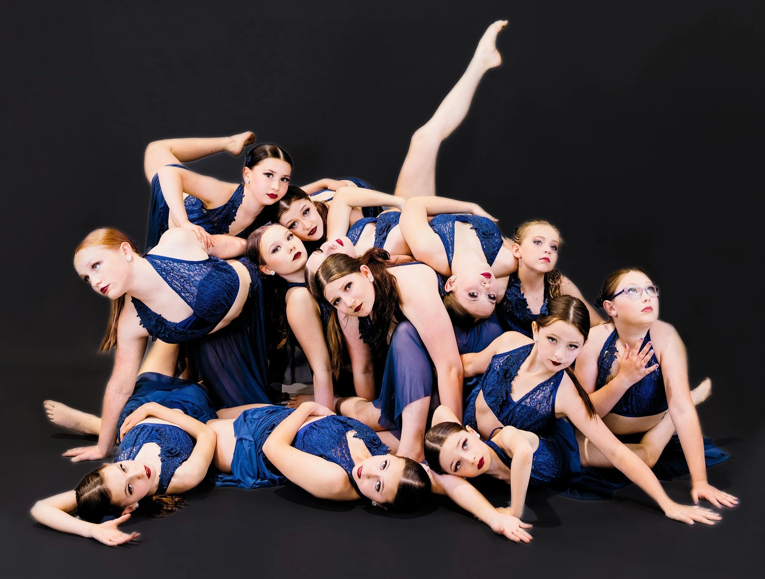 Group of young girls in matching blue dresses and makeup posing on a black background, arranged in a stacked formation with some lying down, sitting, and leaning on each other.