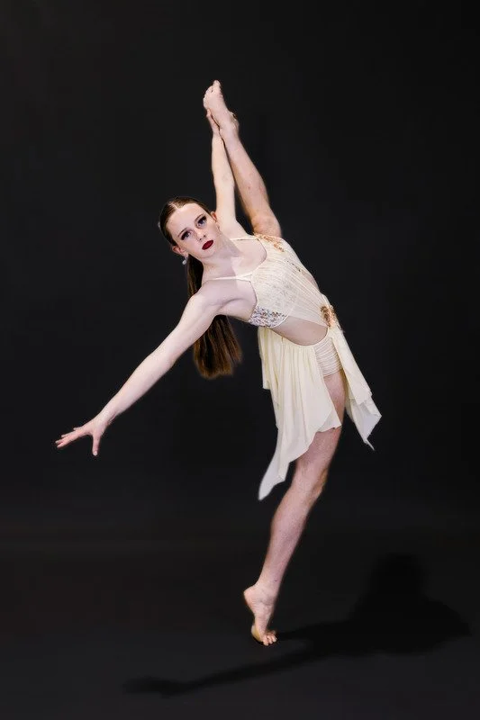 A female ballet dancer in a cream-colored dress performing a pose with one arm raised above her head and the other extended outward, standing on one foot against a black background.