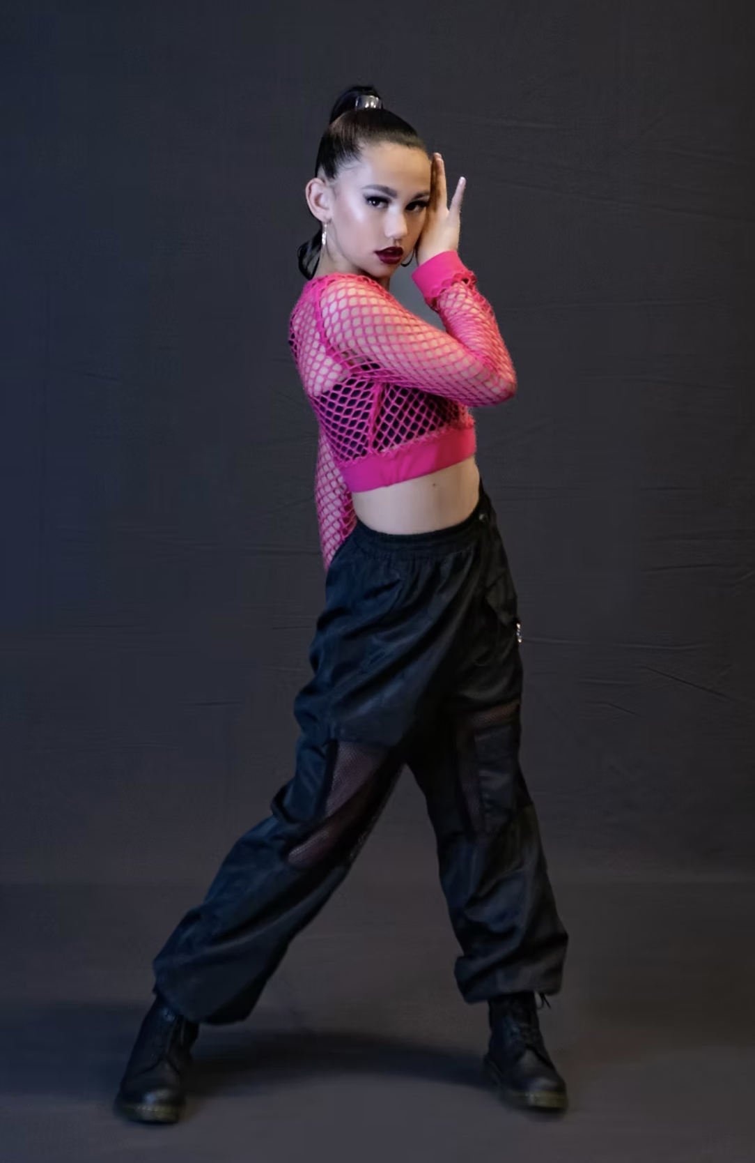 A woman wearing a pink fishnet top, black cargo pants, and black boots, posing against a dark background with her hand on her face and looking at the camera.