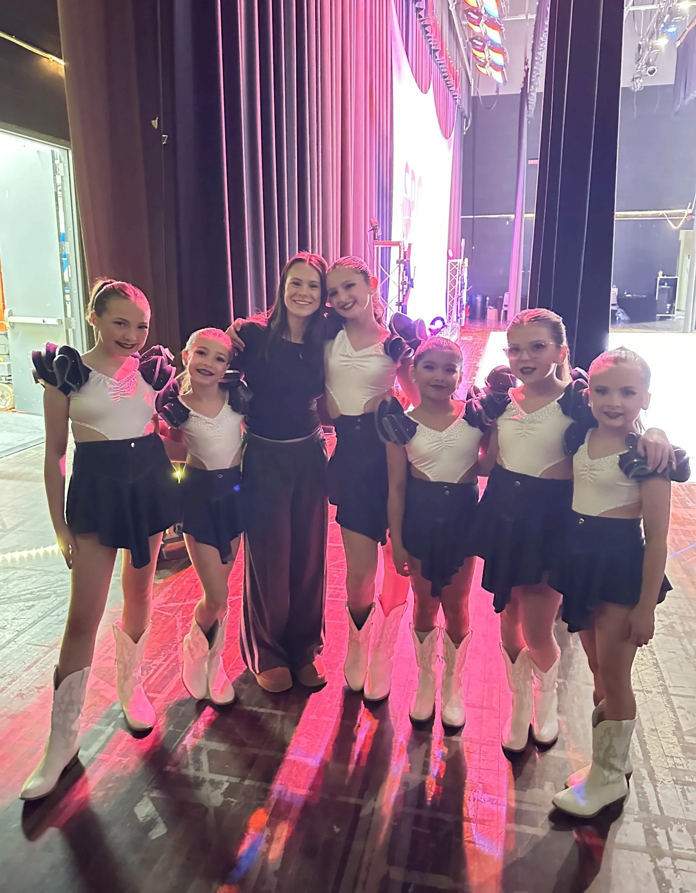 Six young female cheerleaders in black, white, and purple costumes with pom-poms, standing in a backstage area with a stage and curtains behind them, smiling at the camera.