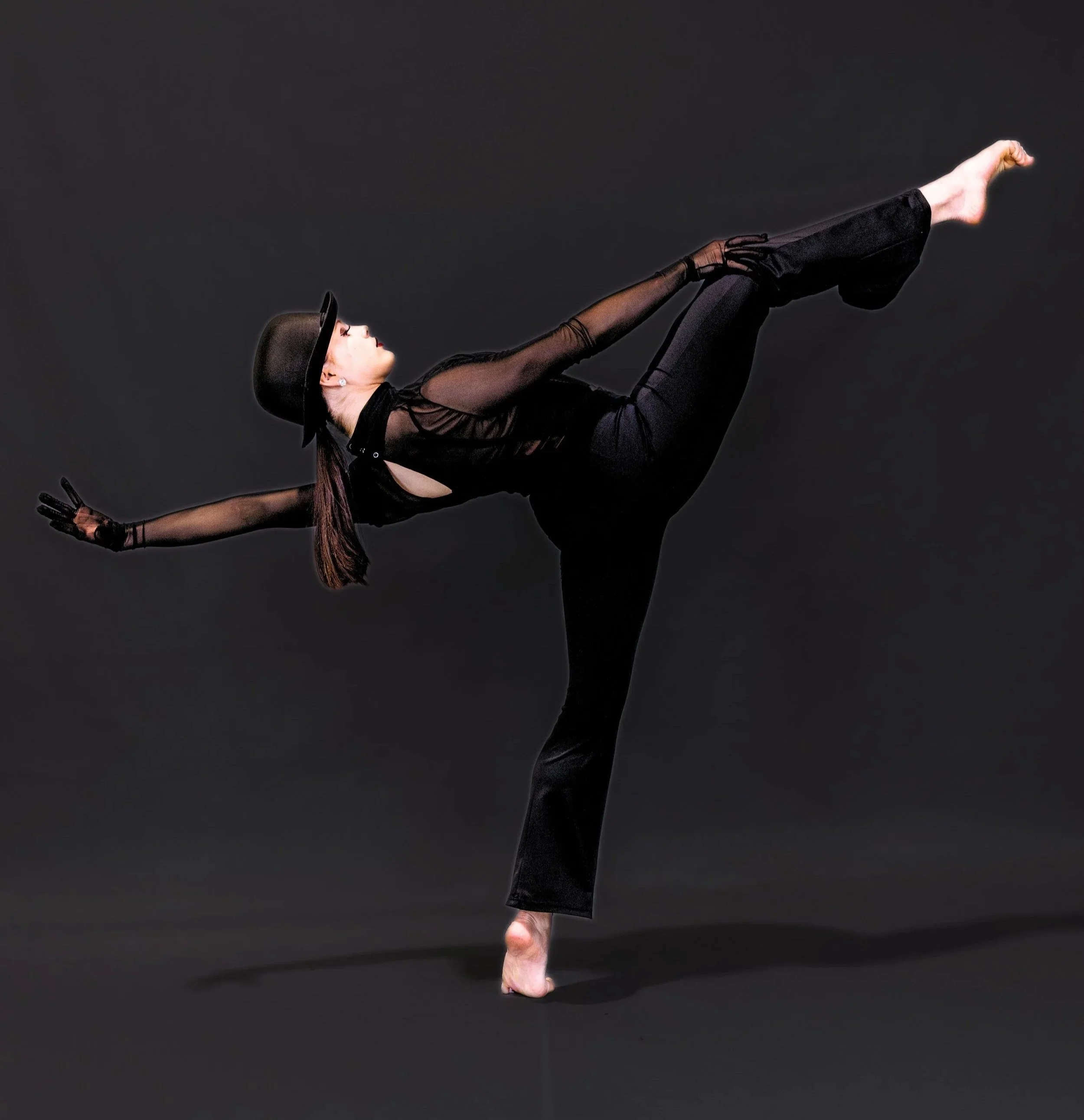 A woman dressed in black, wearing a hat and gloves, is balancing on her finger with her body extended horizontally against a dark background.
