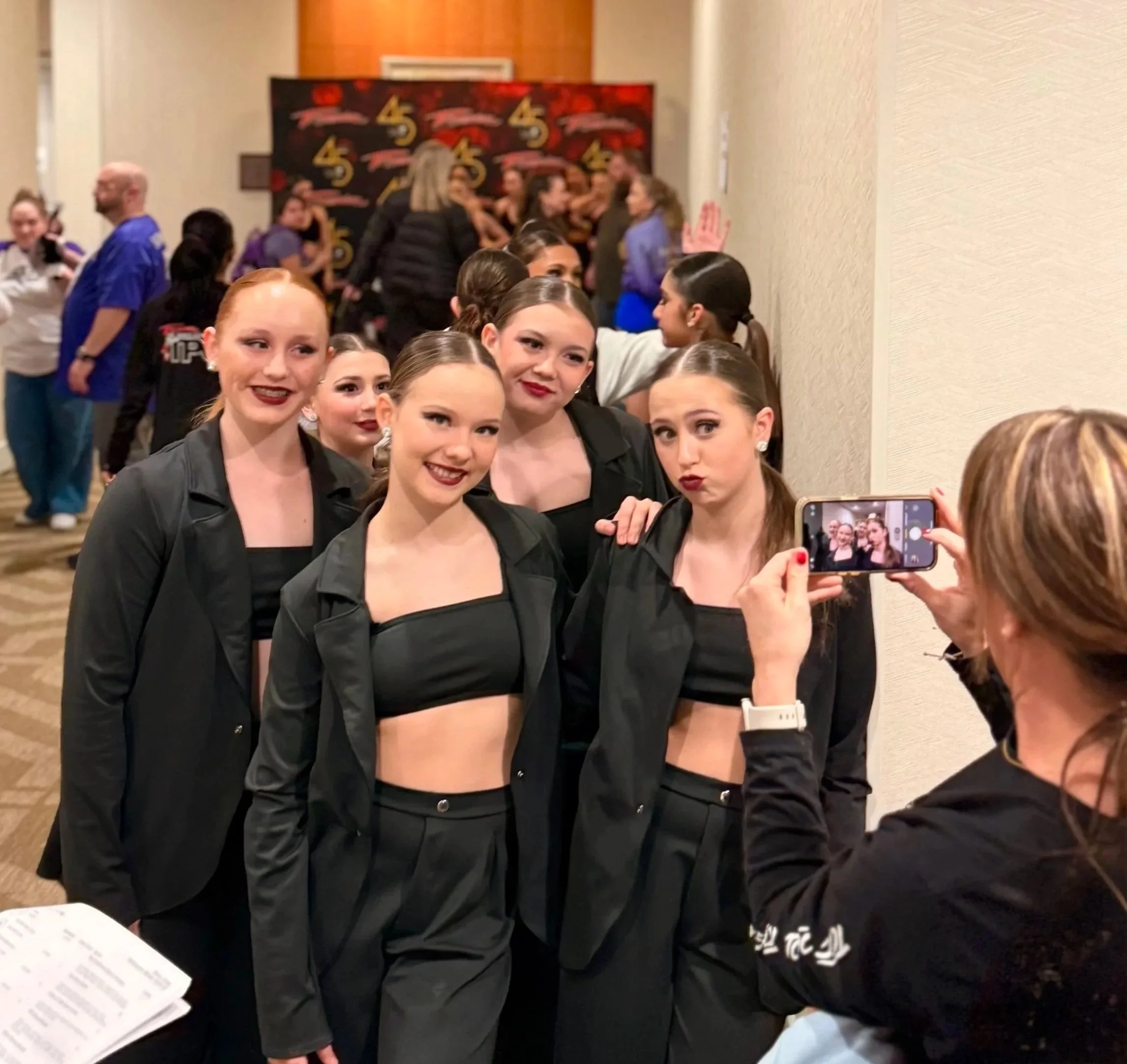 Group of young female dancers in black costumes at a dance competition or event, taking a selfie, with some people in the background and a red and black backdrop.
