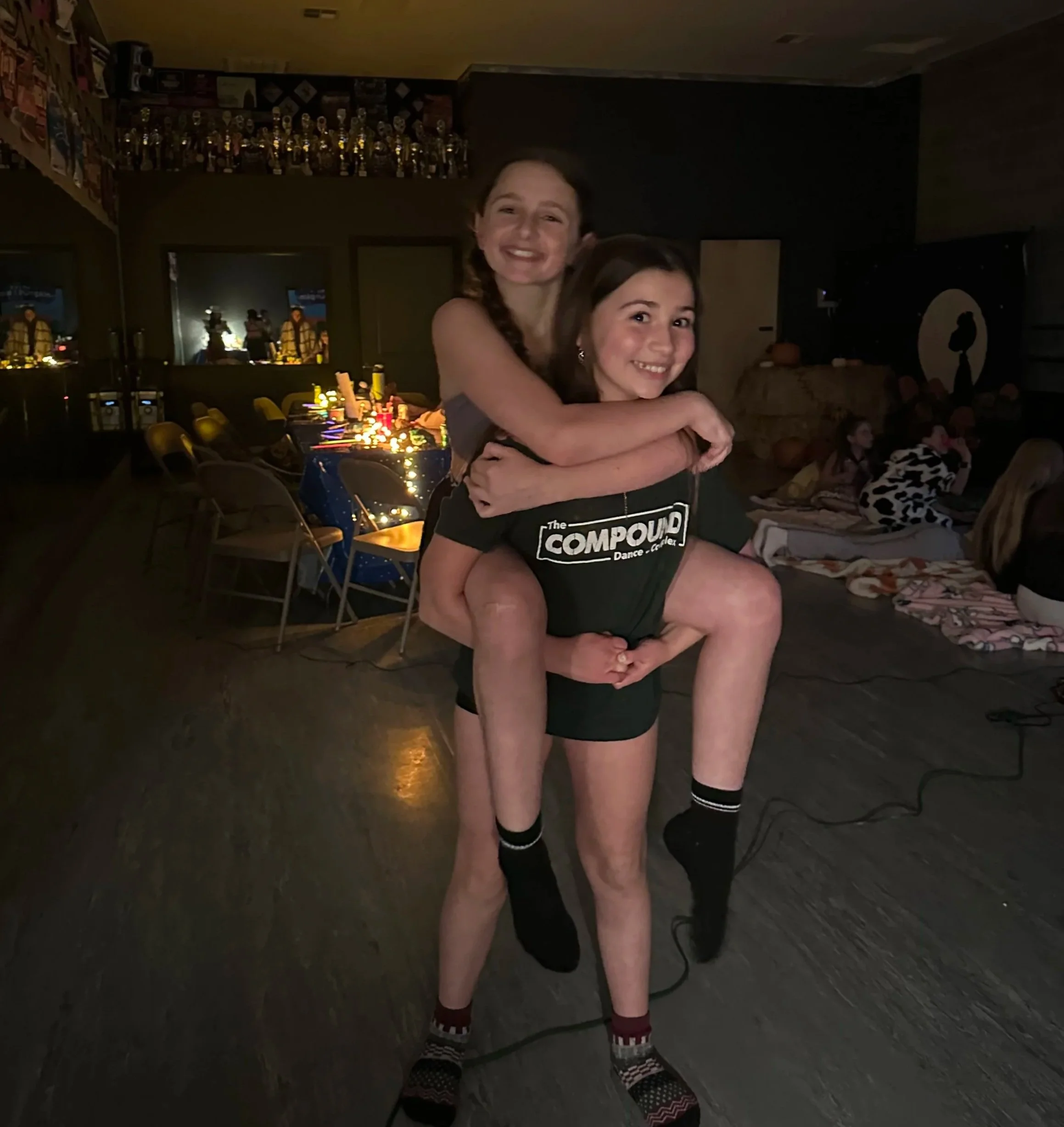 Two young women smiling and hugging each other in a darkly lit indoor setting, with other people sitting on the floor and decorations in the background.