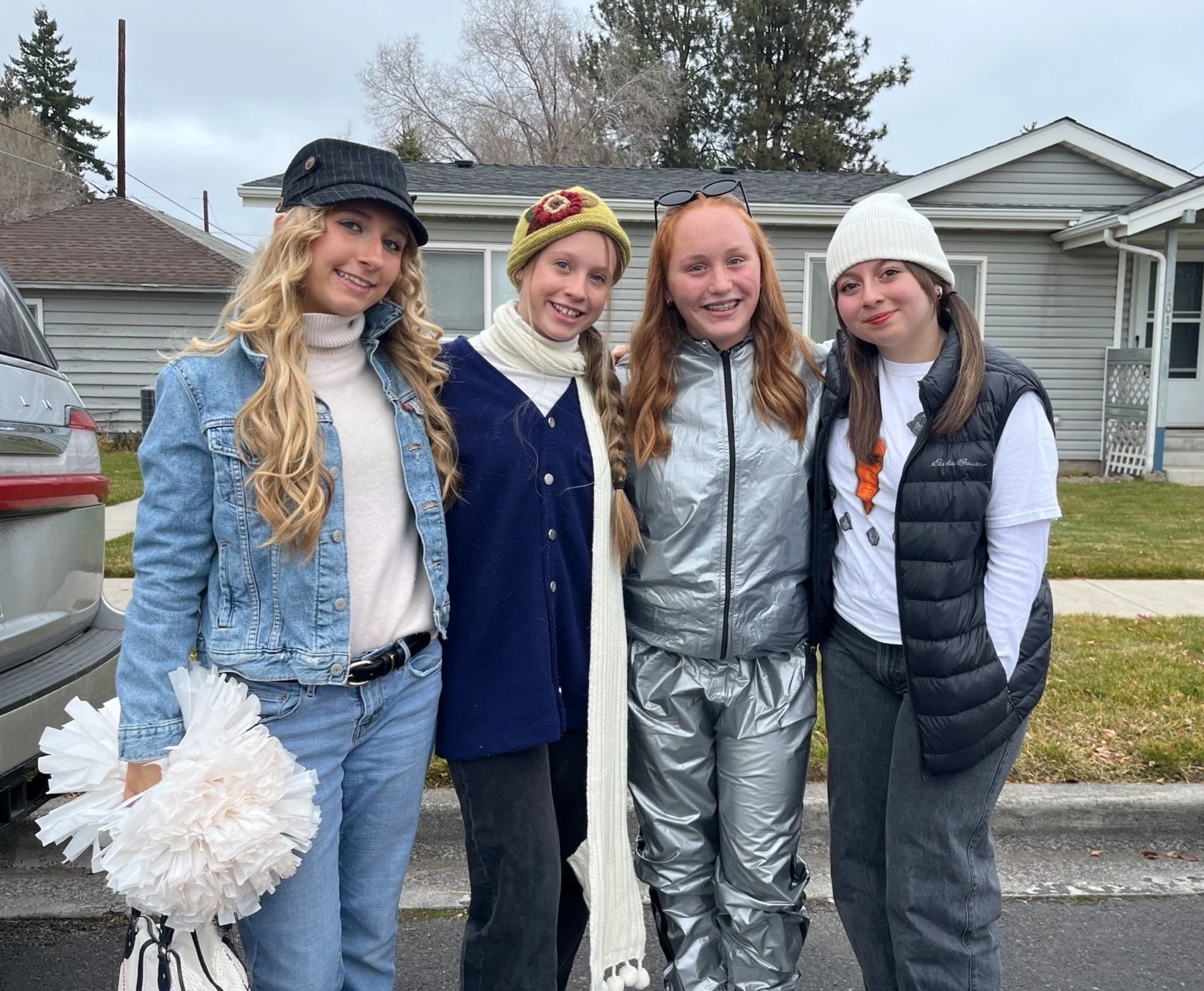 Four young women standing outdoors in front of a house, smiling and posing for the photo. They are dressed in casual, warm clothing including jackets, hats, and scarves, indicating cool weather.