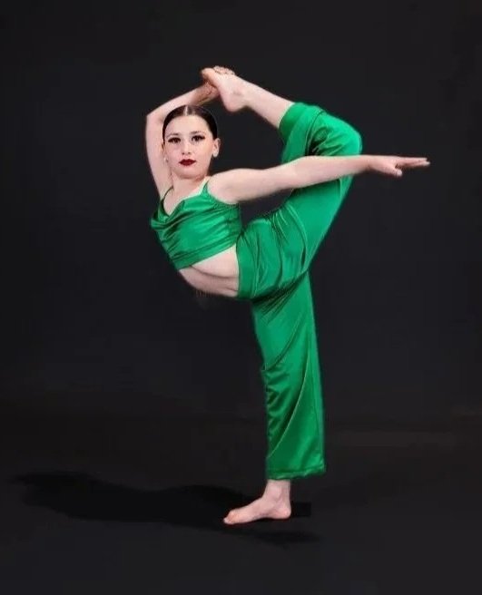 Young woman in green dance costume performing a pose with one leg bent behind her back and one arm stretched forward, against a black background.