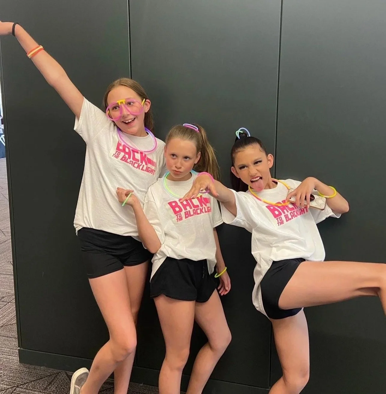 Three young girls are wearing matching white t-shirts with pink text, black shorts, and glow necklaces. They are striking playful poses against a dark gray wall, with one girl making a silly face and the others posing confidently.