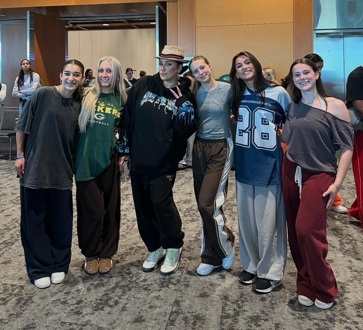 Six young women standing together indoors, smiling and posing for a photo. They are dressed casually in athletic or comfortable clothing, with a wooden wall and other people visible in the background.