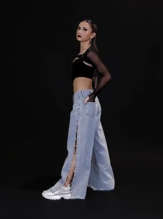 A young woman with long dark hair and makeup, wearing a black crop top with sheer long sleeves, loose light blue jeans with side slits, and white sneakers, standing against a black background.