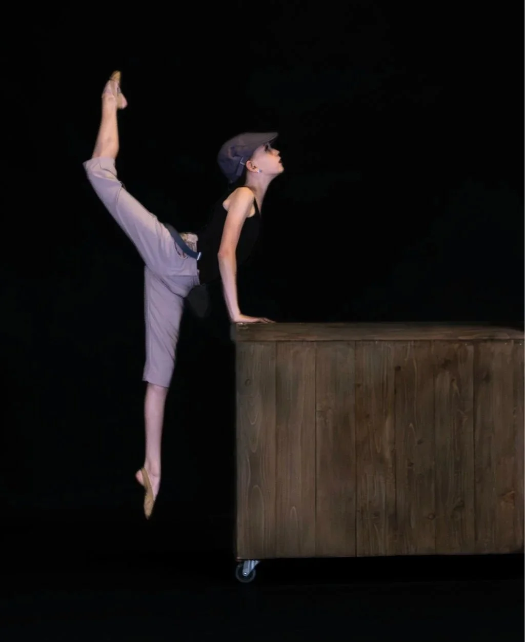 A woman in a black top, beige pants, and a cap is performing a handstand with her leg extended backward in a dance or gymnastics pose, supporting herself with her hands on a wooden block against a black background.