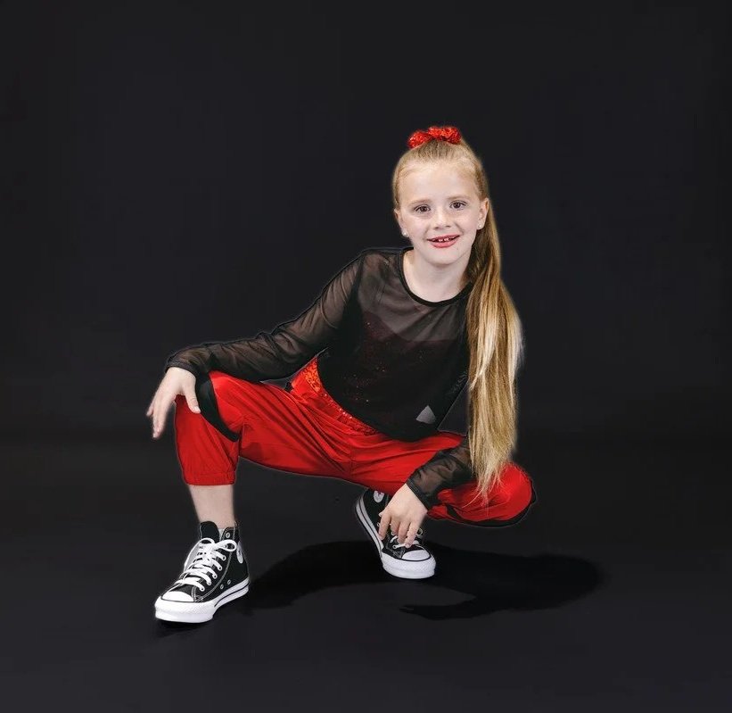 Young girl with long blonde hair, red top bun, squatting in a black and red dance outfit with sneakers, smiling at the camera against a black background.