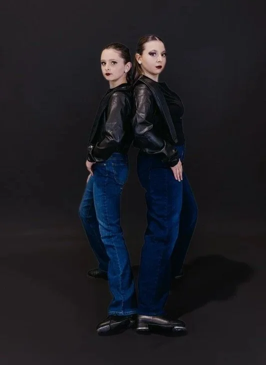 Two women with dark lipstick and makeup, standing back-to-back with their sides touching, wearing black leather jackets and blue jeans, against a dark background.