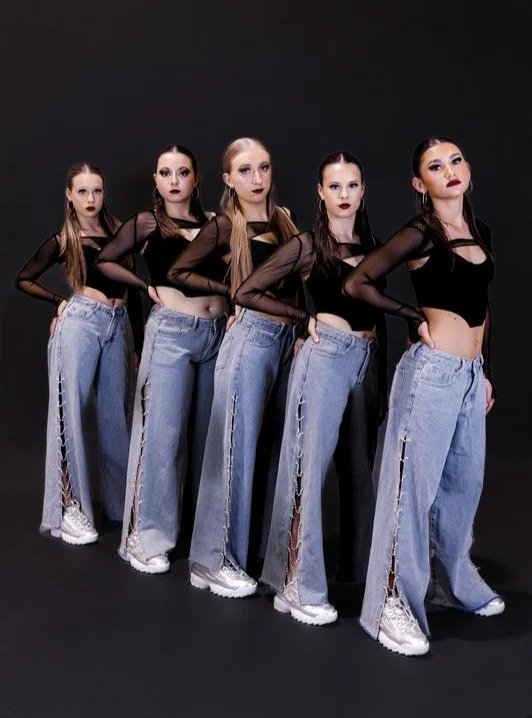 Five young women standing in a line against a dark background, wearing black crop tops and high-waisted jeans with side slits, and white sneakers, posing with confident expressions.