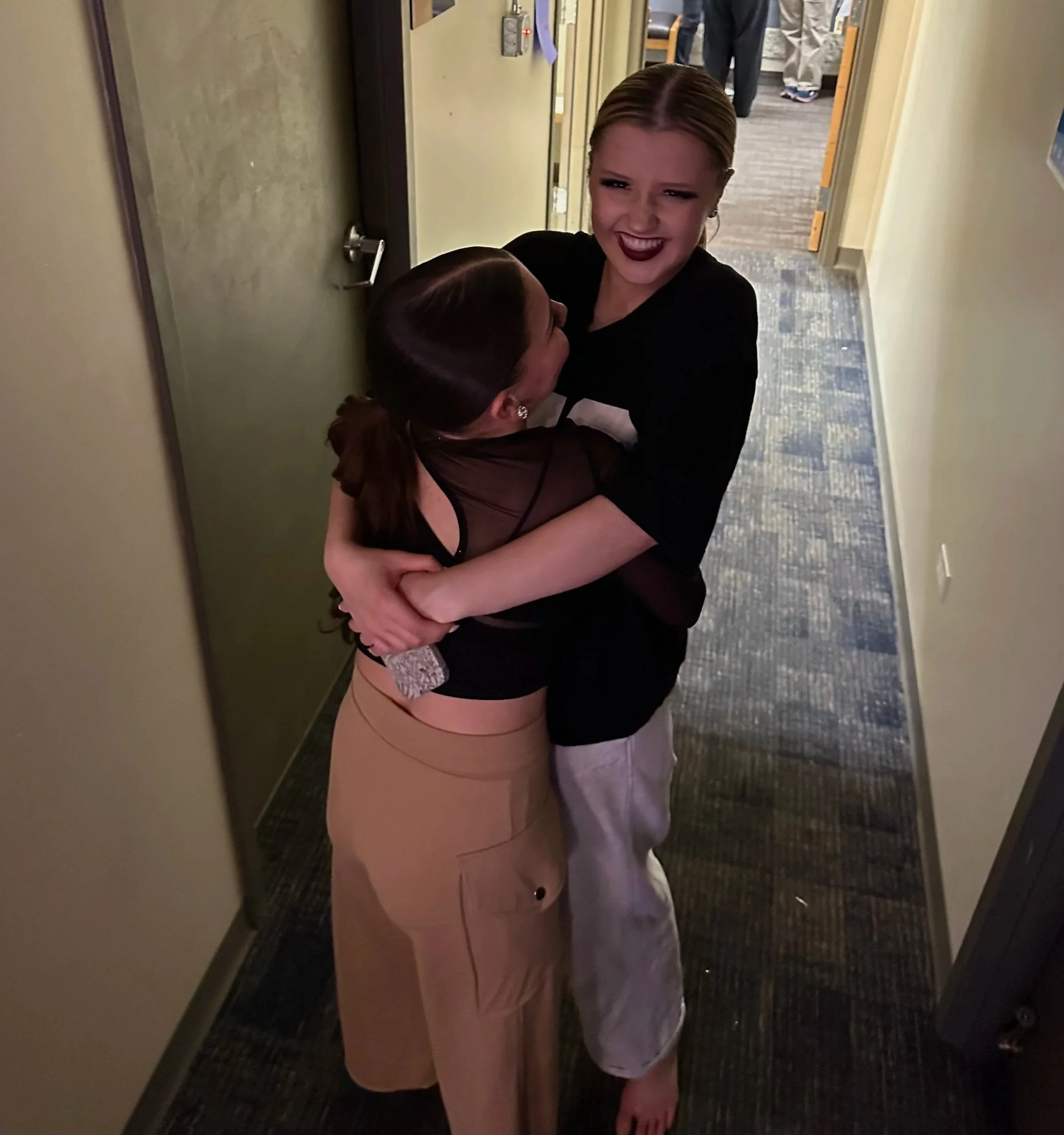 Two women embracing in a hallway, one with short blonde hair and makeup, the other with long brown hair wearing a black dress. The woman with blonde hair is smiling.