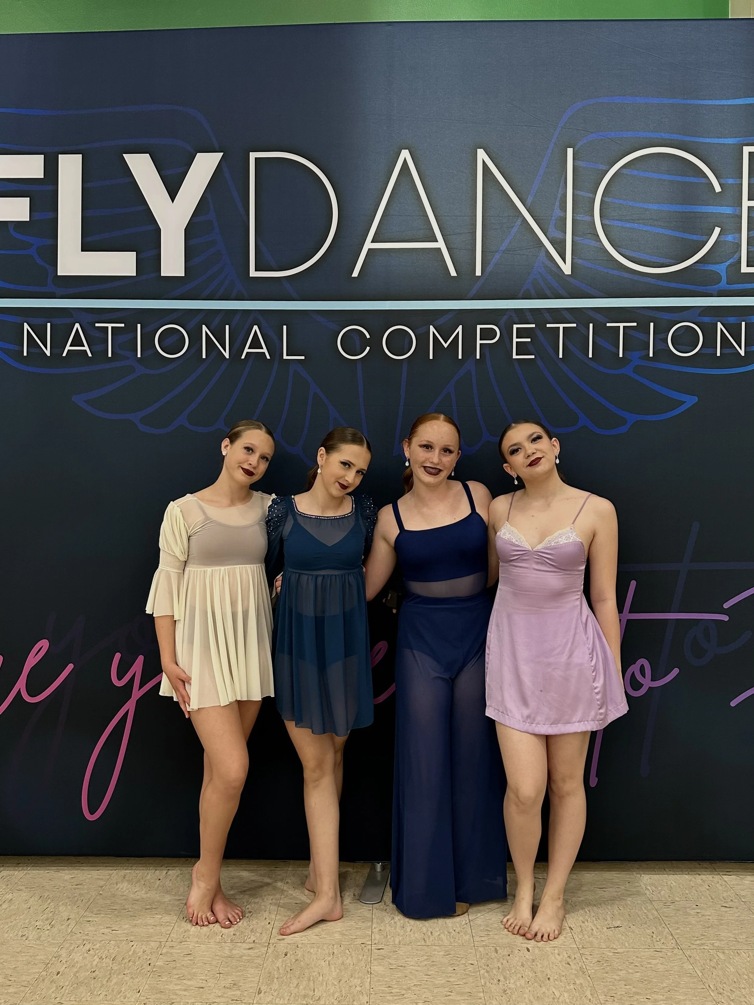 Four young women dressed in dance costumes standing together in front of a large sign that reads 'FlexDance National Competition.'