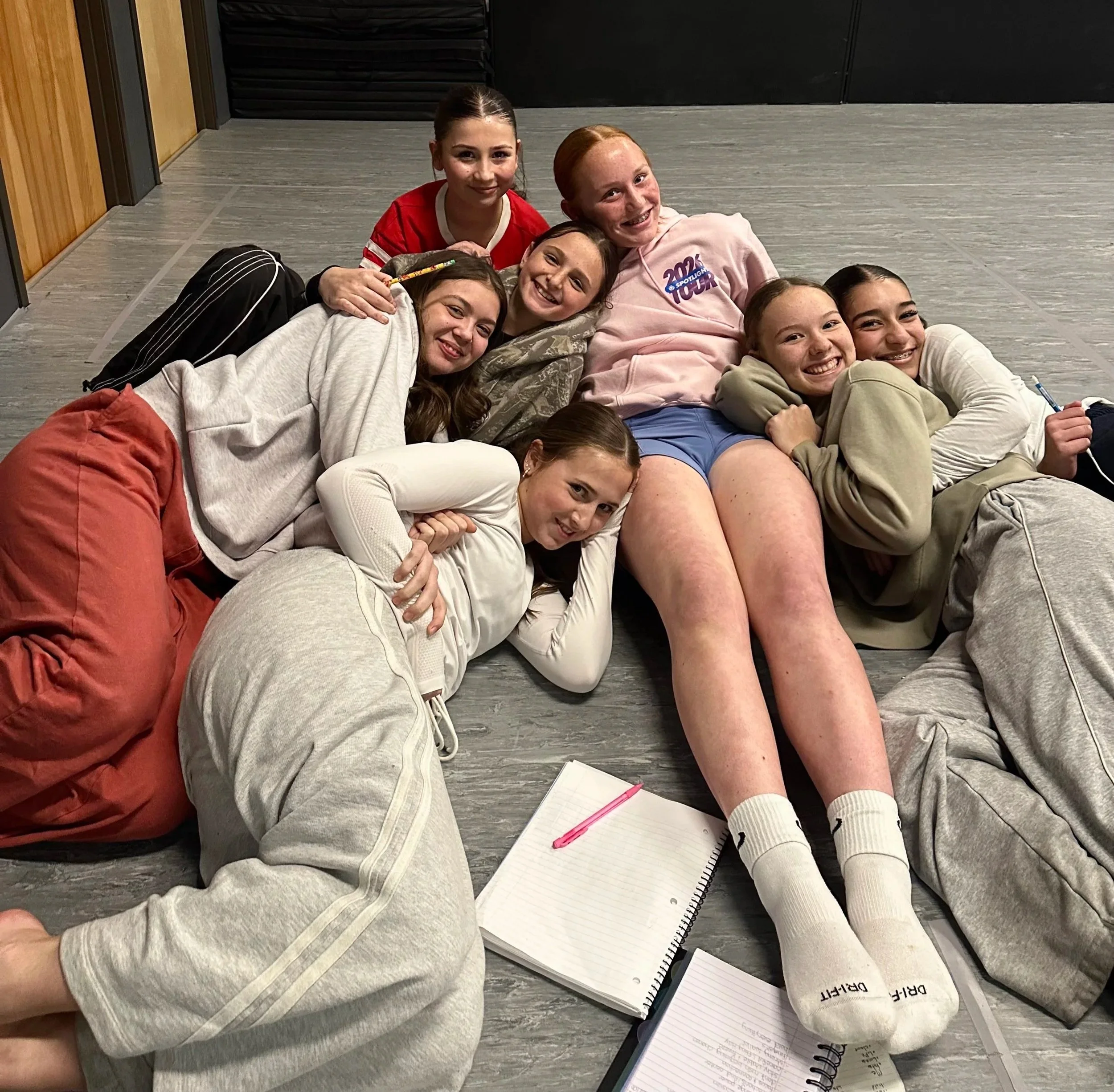 Six girls lying and sitting on the floor together, smiling and posing for a group photo. They have various hair colors and styles and are dressed casually. There are notebooks and pens on the floor nearby.