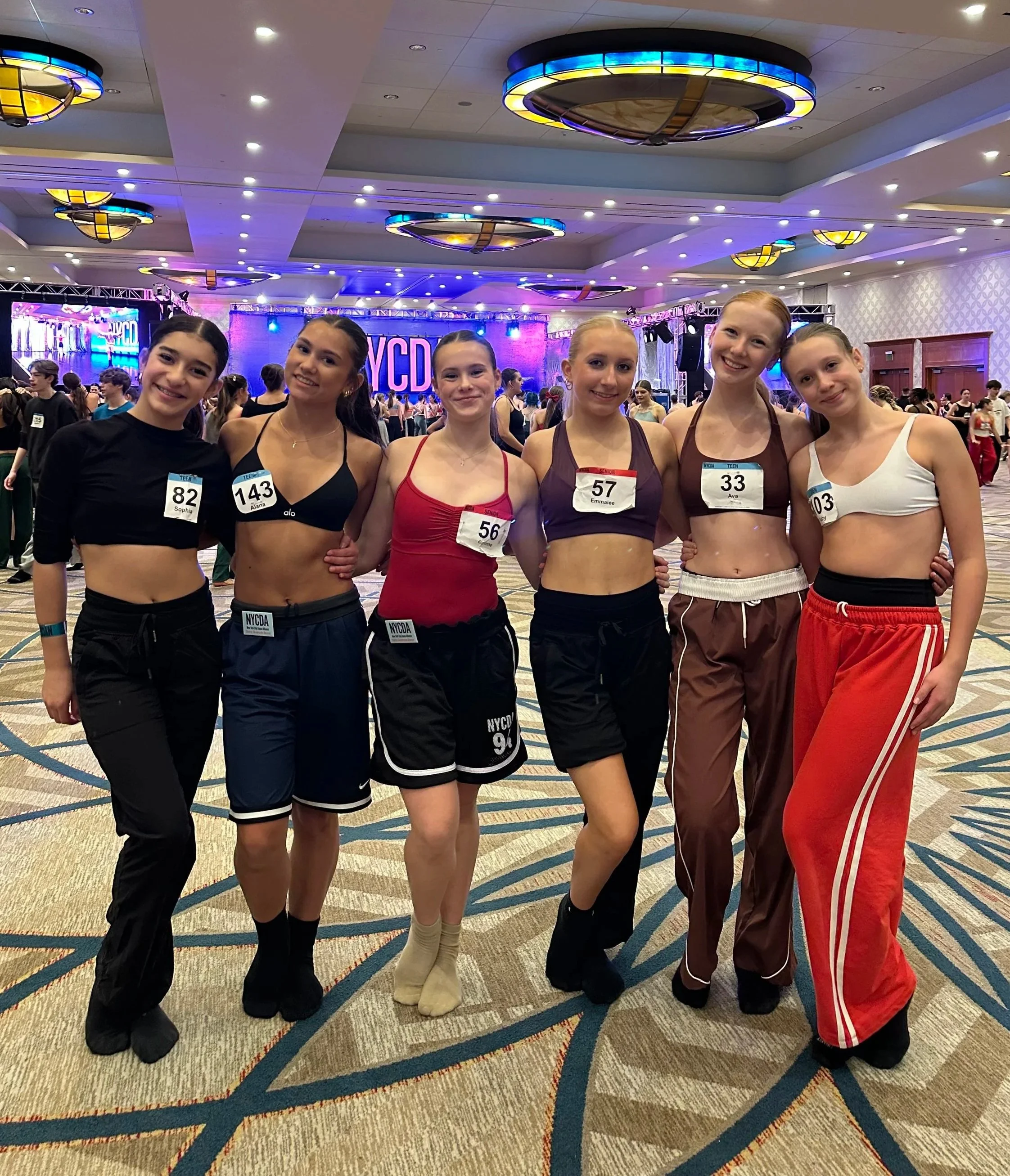 A group of six young women standing in a ballroom at a dance competition, smiling at the camera, wearing athletic outfits and competition badges, with a stage and large screen displaying 'NYCDA' in the background.