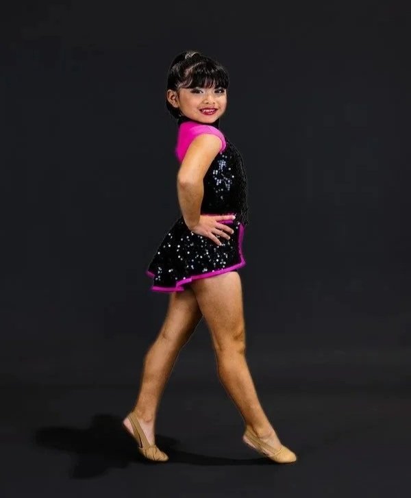 A young girl in a colorful dance outfit, standing in a confident pose with one hand on her hip, smiling against a dark background.
