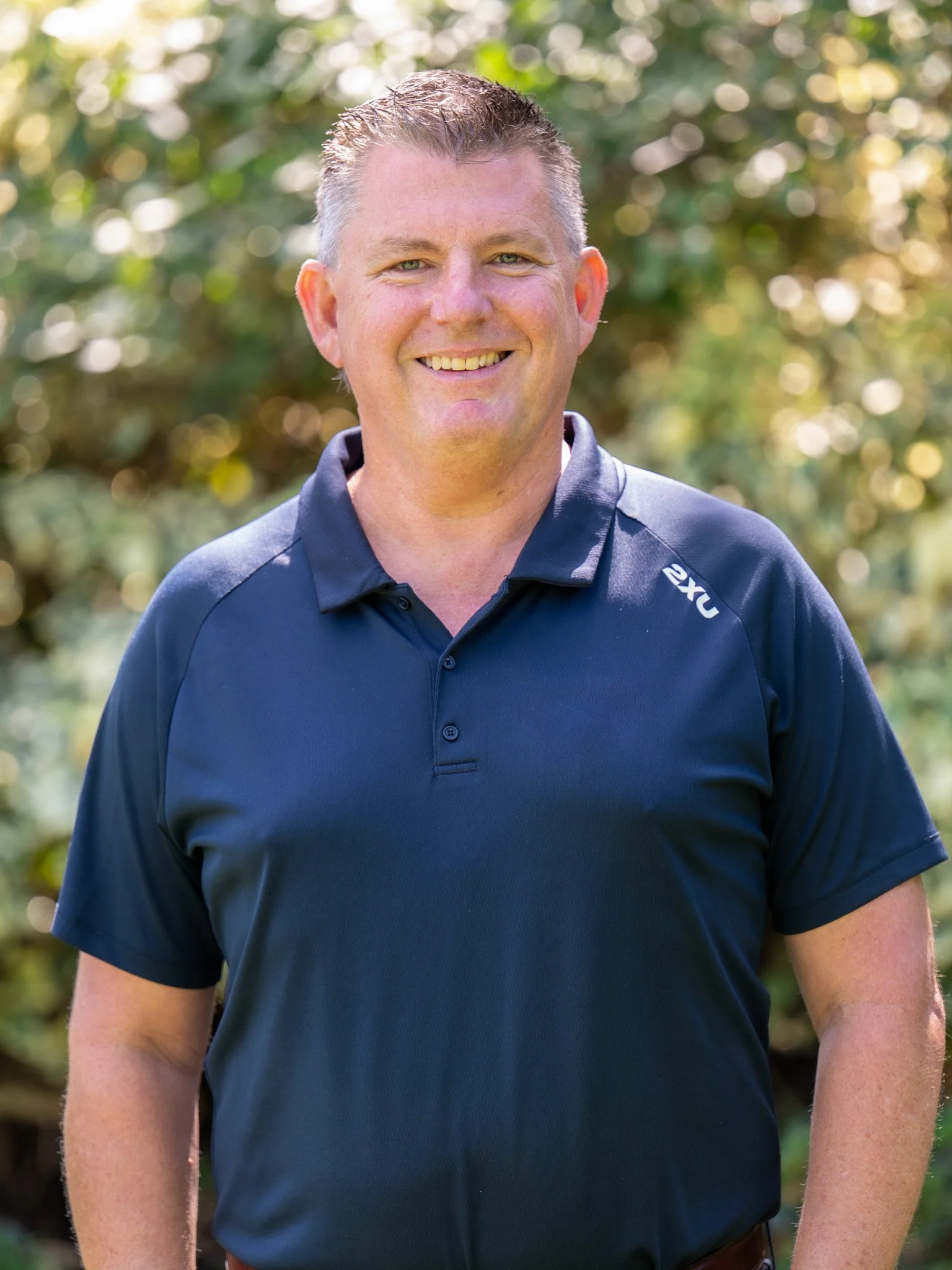 Bunbury Physio - A smiling man with short hair standing outdoors with a blurred, leafy background.