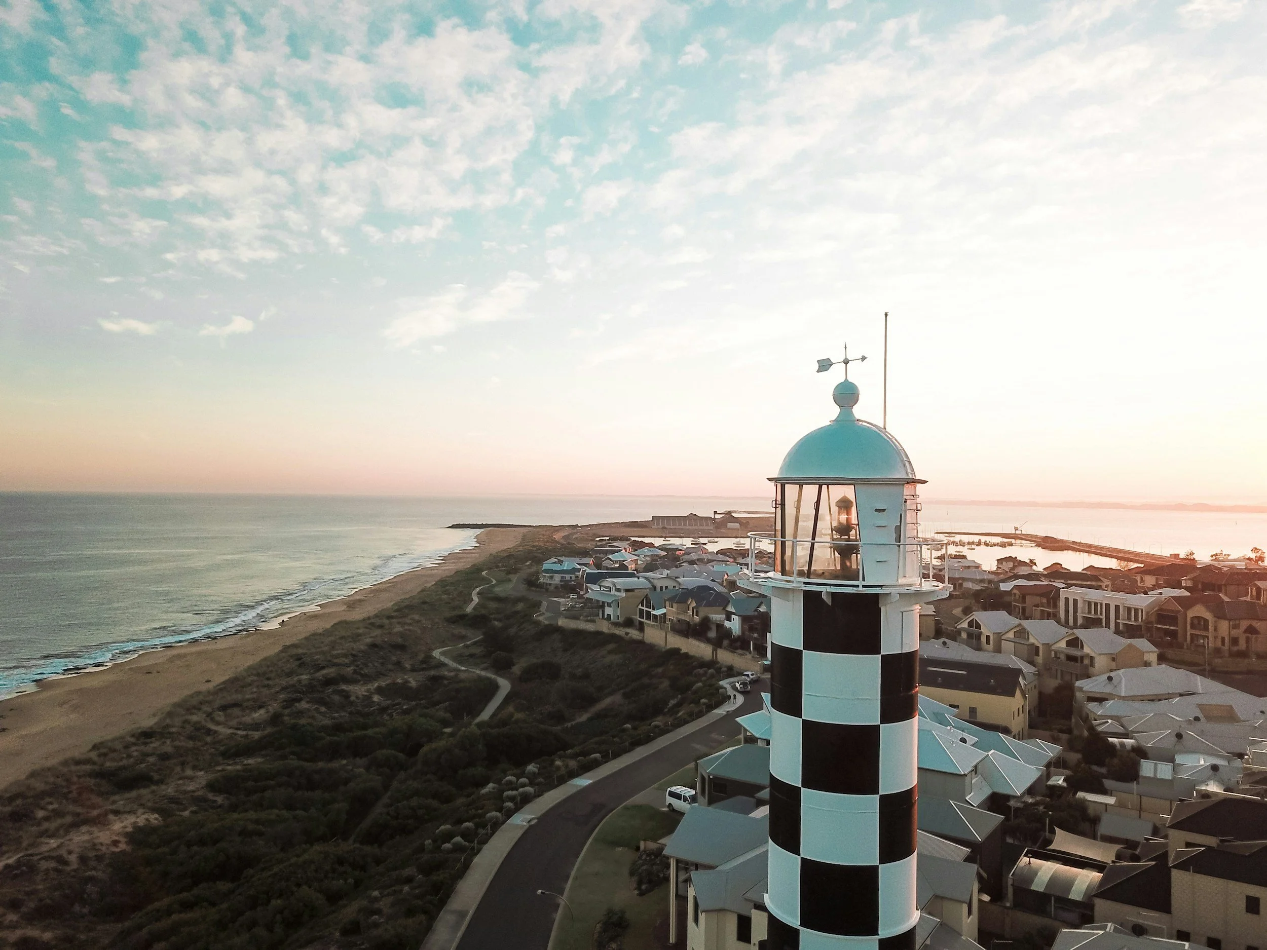Bunbury Physio - A coastal scene at sunset with a distinctive black and white checkered lighthouse in the foreground, overlooking houses, the ocean, and a cloudy sky.