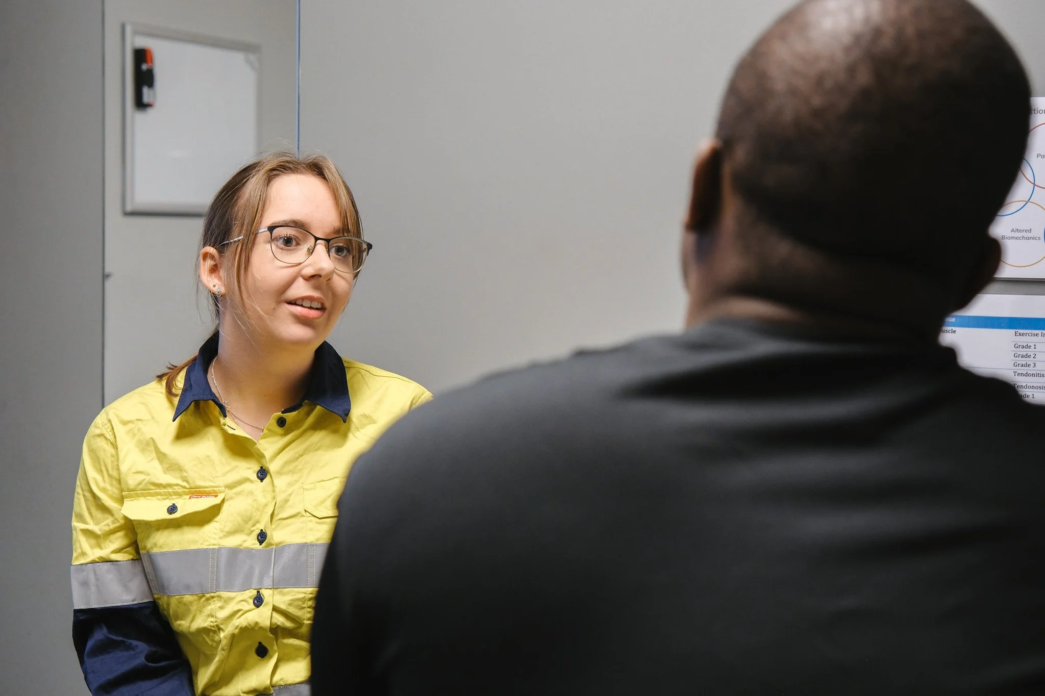 Bunbury Physio - A young woman with glasses and a yellow safety jacket talking to a man in a dark suit, in front of a mirror in an office or medical setting.