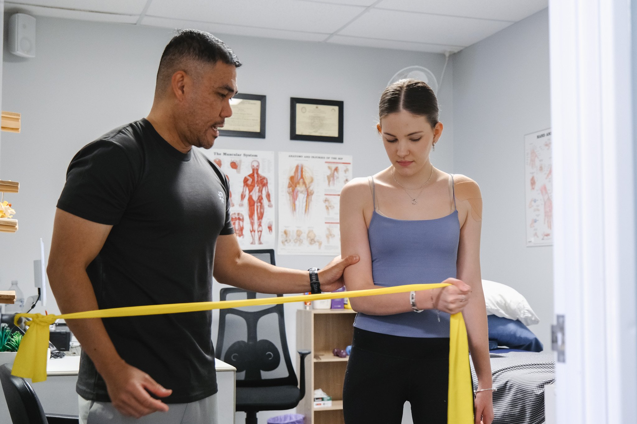 Bunbury Physio - A man explaining something to a young woman in a medical setting, with charts of human anatomy on the wall, and a yellow caution tape in the foreground.