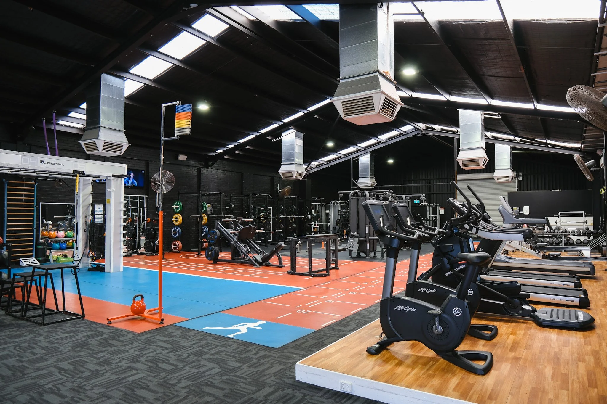 Bunbury Physio - Interior of a modern gym with exercise equipment, including treadmills, weightlifting stations, and free weights. The ceiling has large vents and skylights, with a mix of dark and light flooring areas.