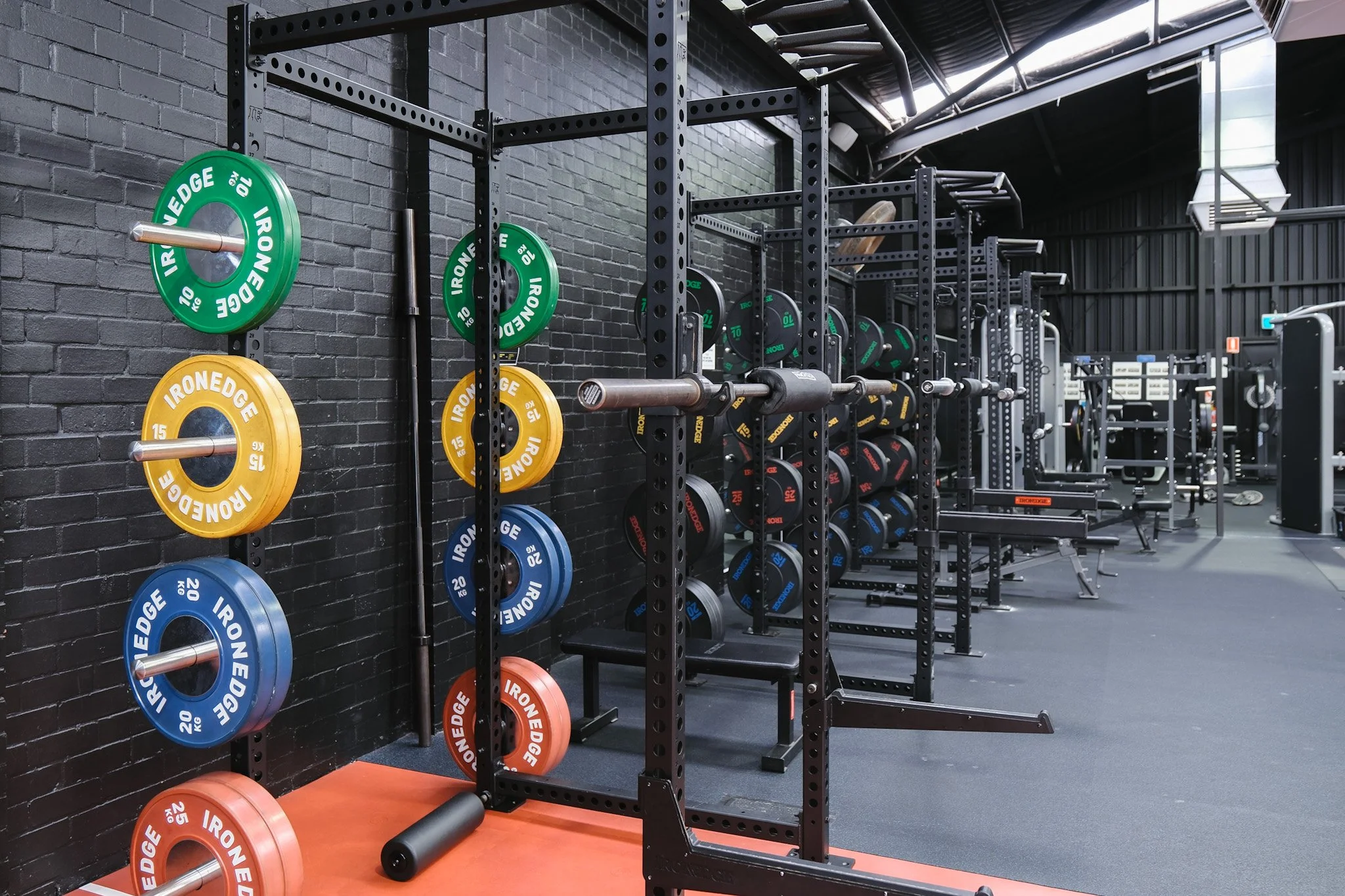 Bunbury Physio - Exercise gym with colorful weight plates and black metal squat racks, black brick wall background, and gray flooring.