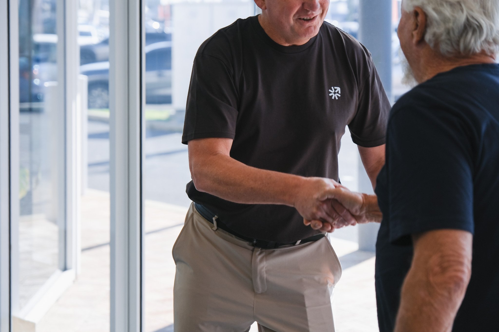 Bunbury Physio - Two men are shaking hands inside a building near glass doors, smiling at each other. One man is wearing a black T-shirt, and the other is wearing a dark-colored shirt. The background shows a parking lot and some cars outside.