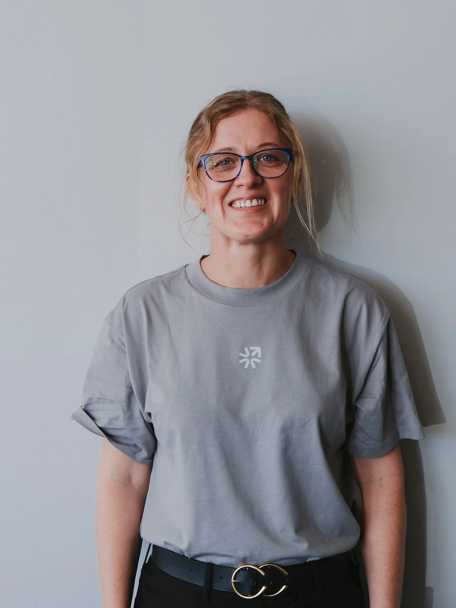 Bunbury Physio - A woman with curly hair, glasses, and light skin, smiling, stands against a plain light-colored wall. She is wearing a gray t-shirt and black pants with a black belt featuring a double G gold buckle.
