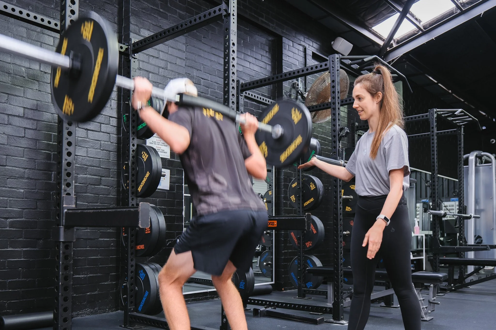 Bunbury Physio - A woman spotting a man lifting weights in a gym with black brick walls and gym equipment in the background.