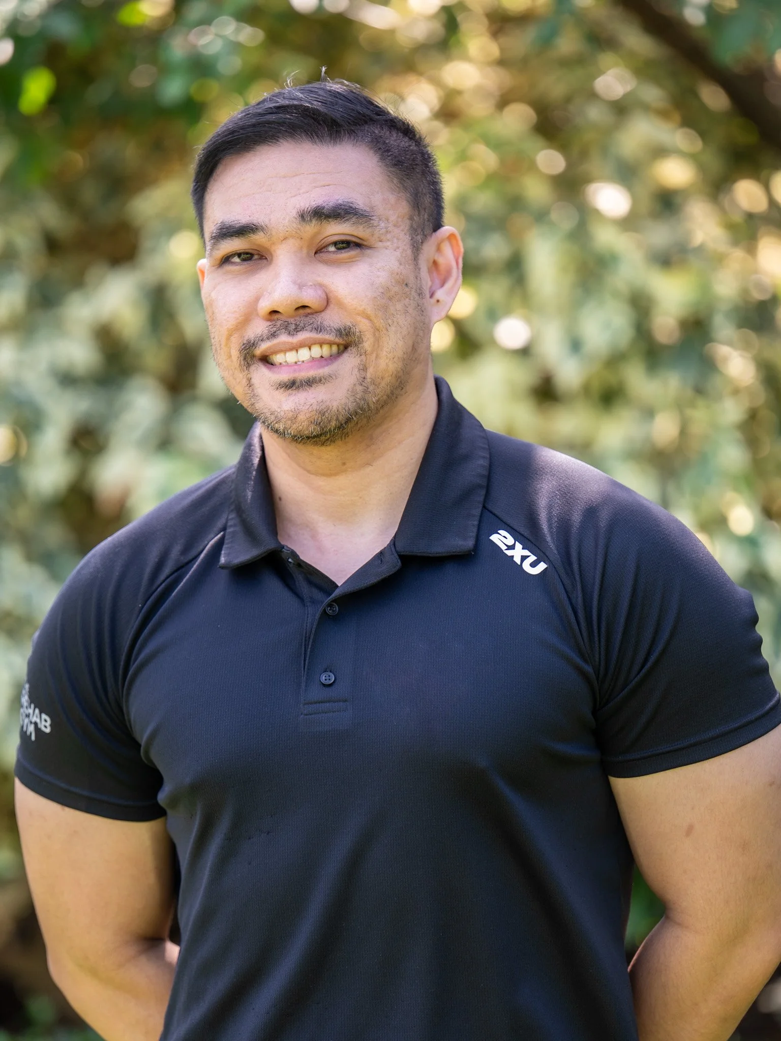Bunbury Physio - Close-up of a man with short dark hair, a light beard, and tan skin, smiling outdoors with a blurred green background.