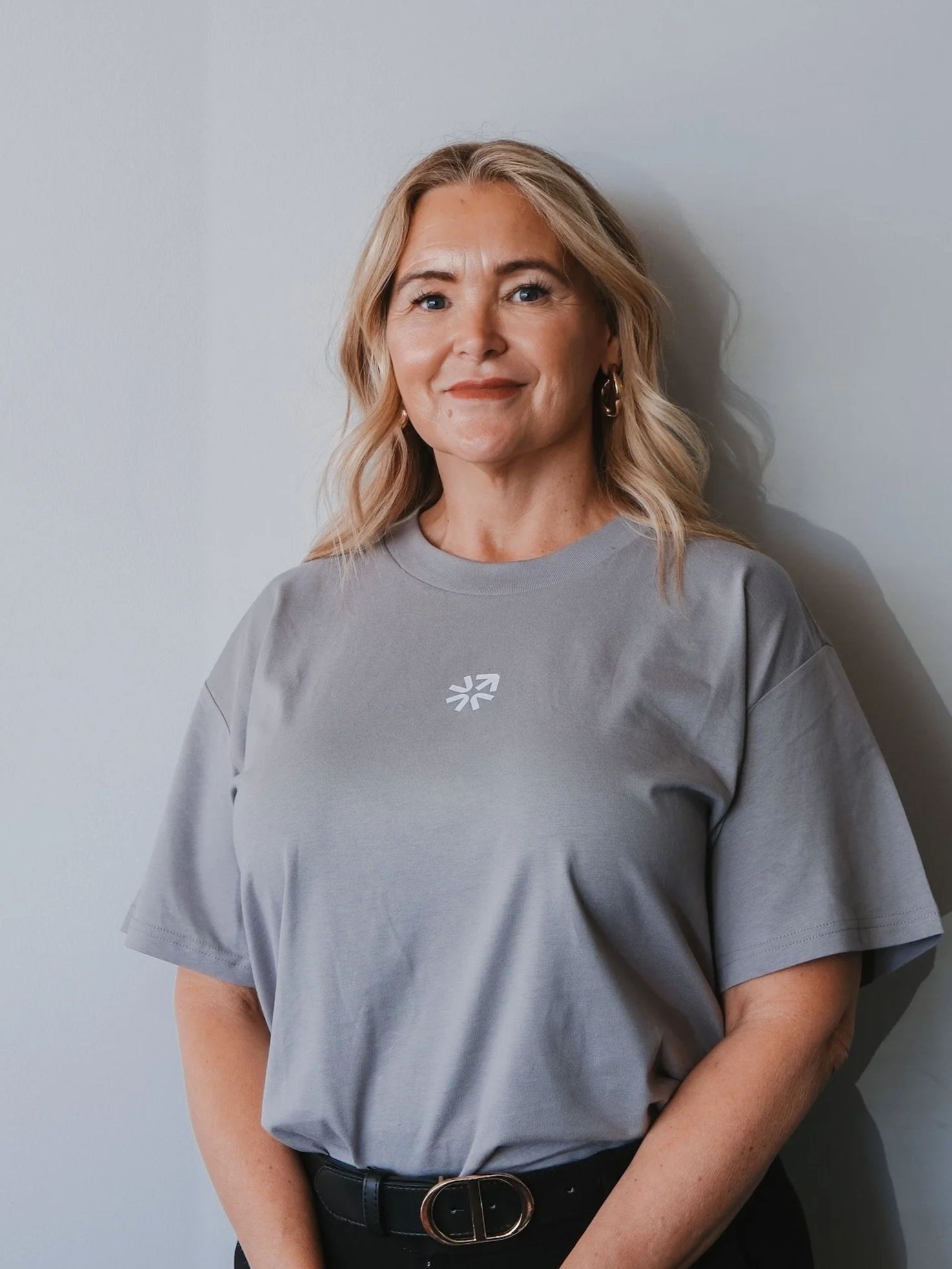 Bunbury Physio - A woman with blonde hair, blue eyes, wearing a gray T-shirt and black belt, standing against a plain gray wall.