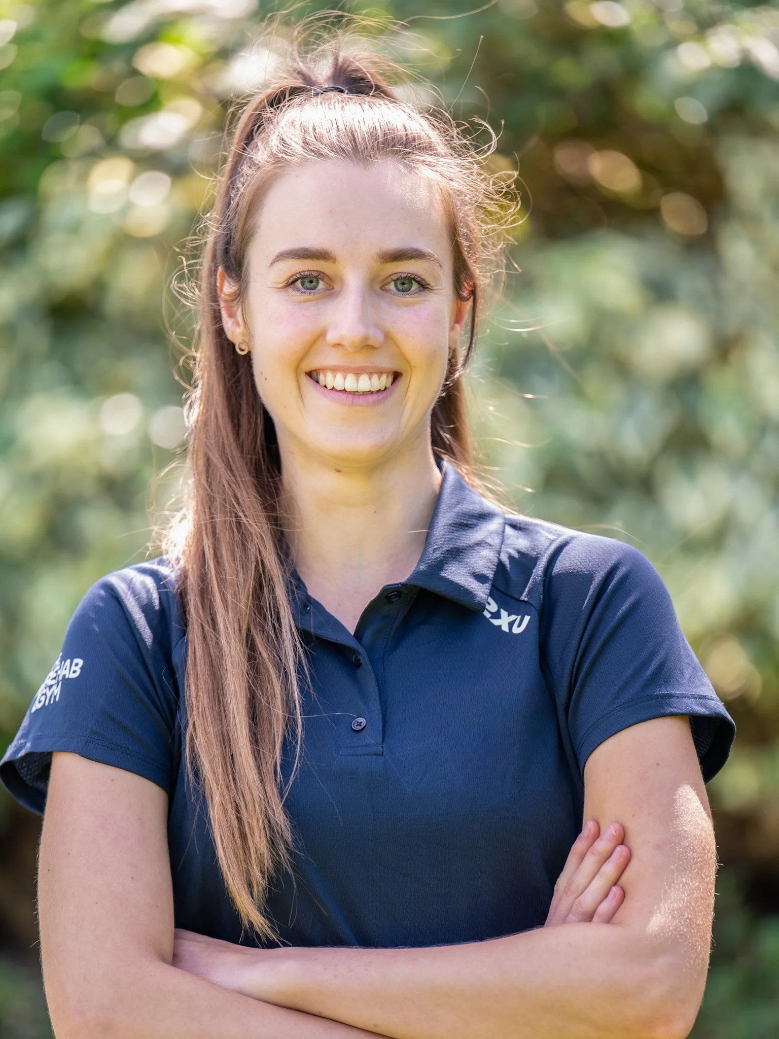 Bunbury Physio - Young woman with long brown hair tied back, wearing a navy sports shirt, standing outdoors with her arms crossed, smiling at the camera, with a blurred green leafy background.