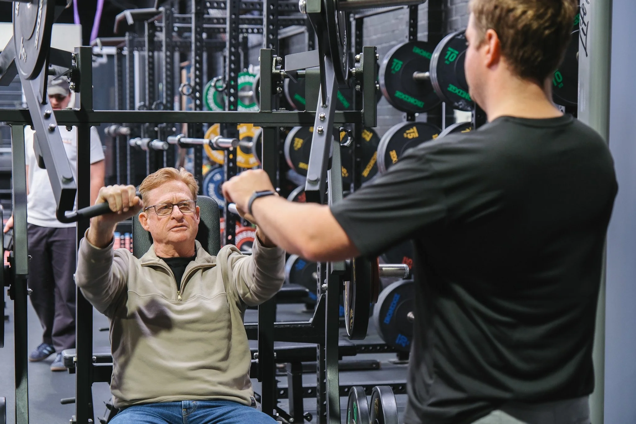 Bunbury Physio - A man performing a seated shoulder press exercise on a gym machine with weights, while another person stands nearby, and a third person is visible in the background in a gym setting.