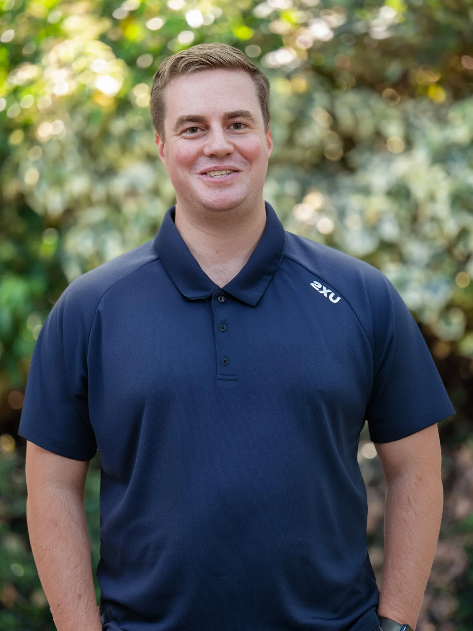 Bunbury Physio - Portrait of a smiling man wearing a navy polo shirt with '2XU' logo, in an outdoor setting with a blurred green and yellow background.