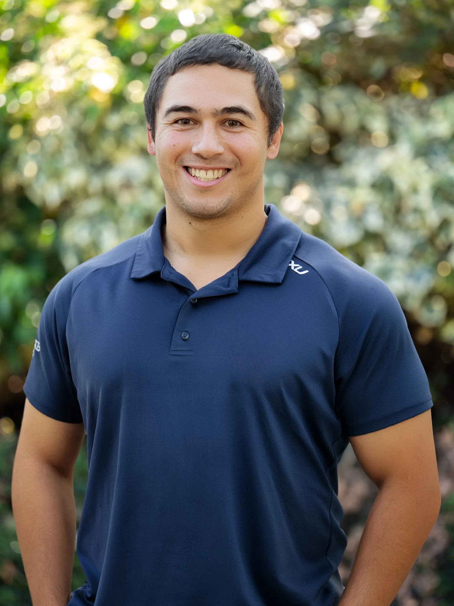 Bunbury Physio - Smiling young man wearing a navy blue polo shirt outdoors with blurred green foliage background.