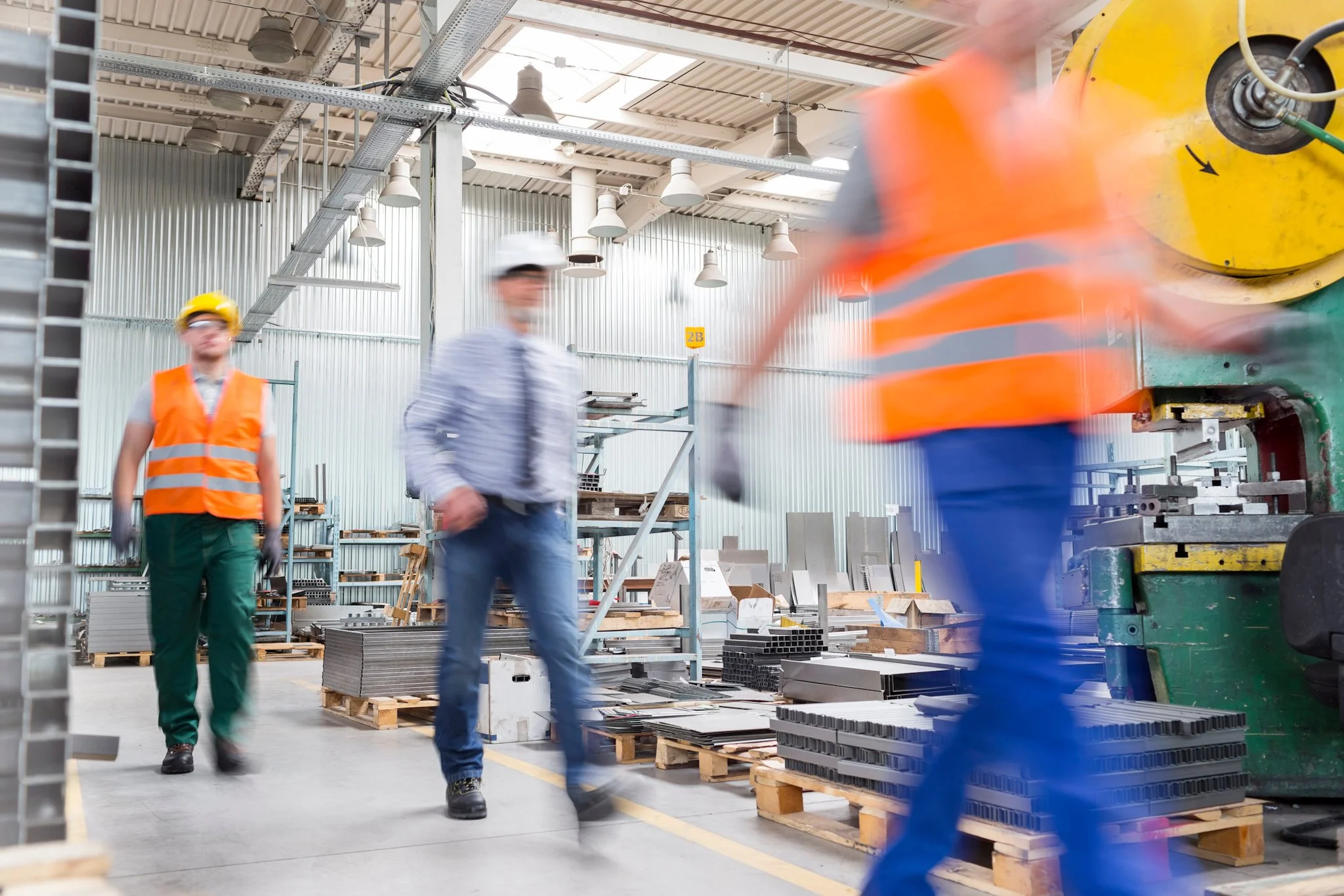 Bunbury Physio - Workers in safety helmets and vests walking and working inside a large industrial warehouse with metal walls, pallets, and machinery.