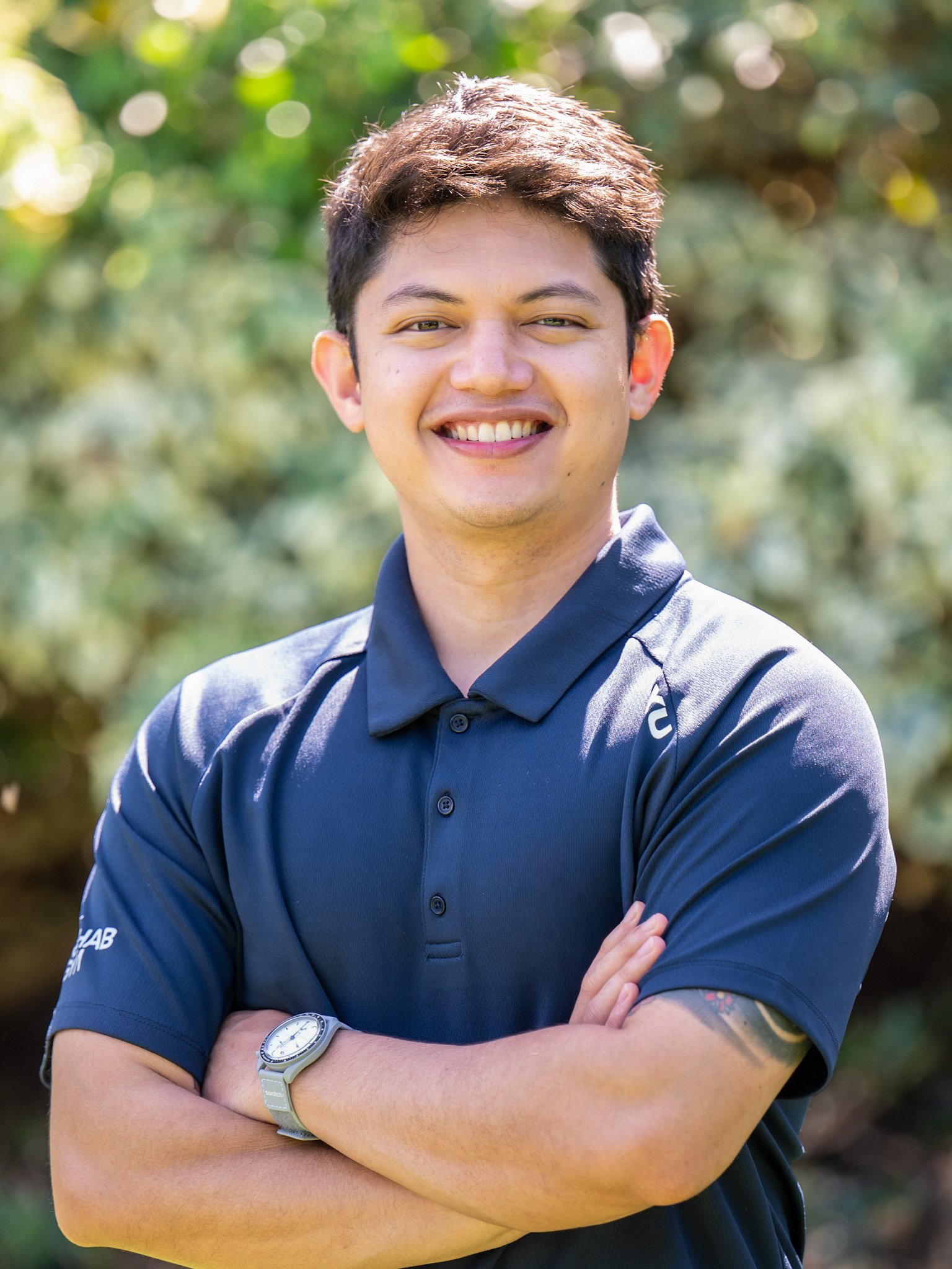 Bunbury Physio - A young man with short brown hair, wearing a navy polo shirt with a logo on the sleeve, standing outdoors with arms crossed and smiling in front of a blurred natural background.