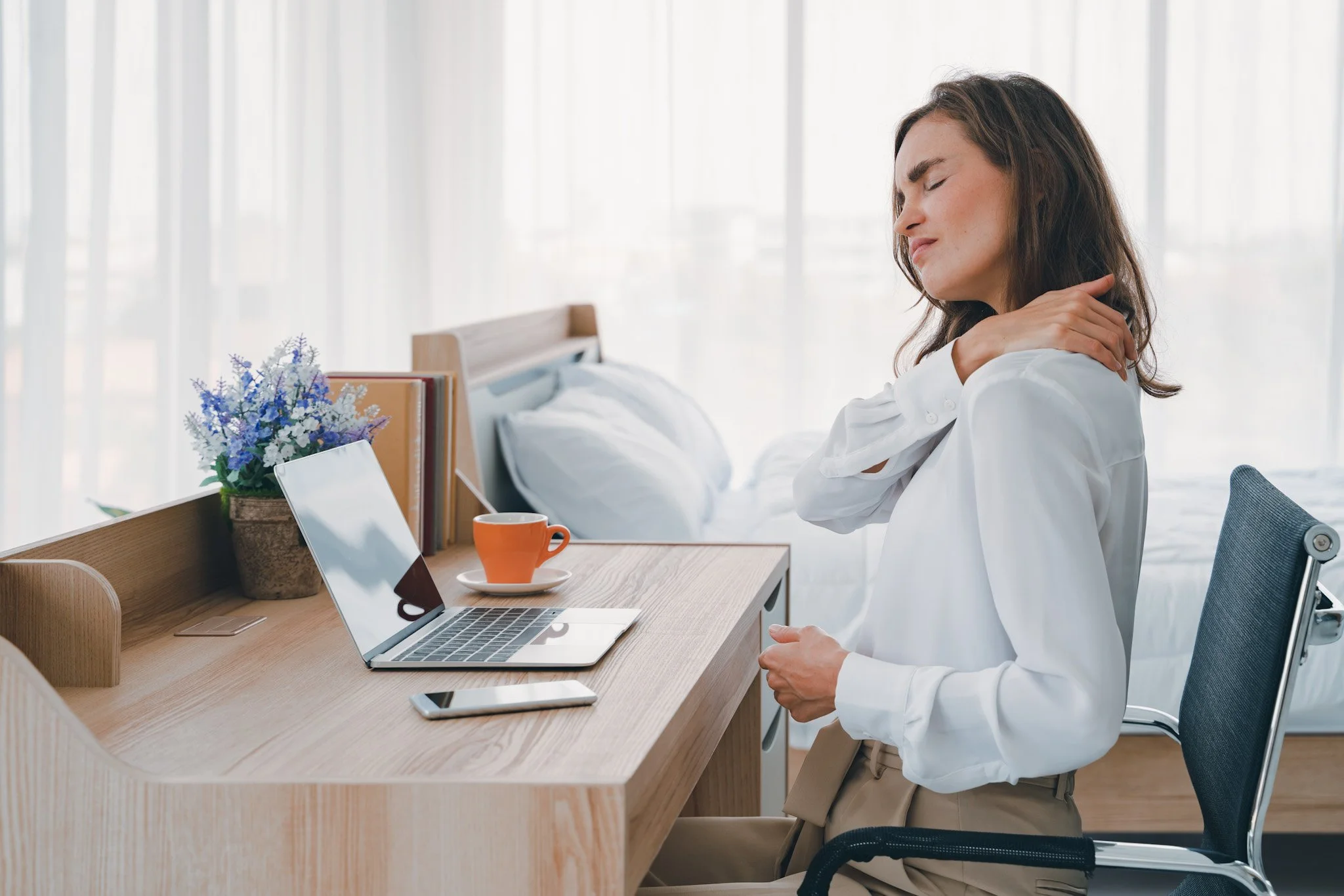 Bunbury Physio - A woman sitting at a desk in a bright room with large windows, stretching or massaging her neck, experiencing neck pain. The desk has a laptop, a tablet, a smartphone, a cup, a flowerpot with purple flowers, and some books.