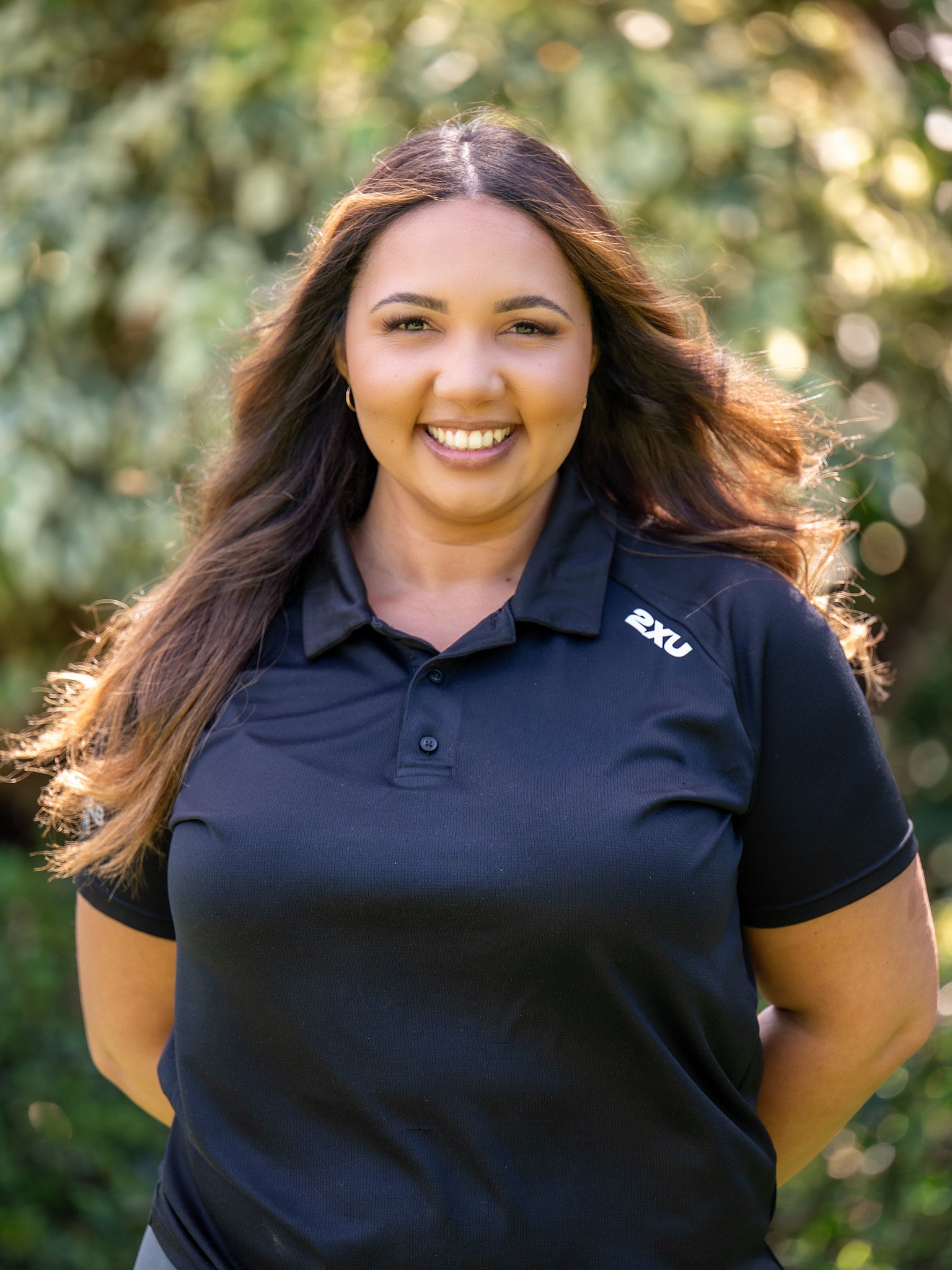 Bunbury Physio - A woman with long brown hair smiling outdoors, wearing a navy blue polo shirt with a logo, in a natural setting with greenery and sunlight in the background.