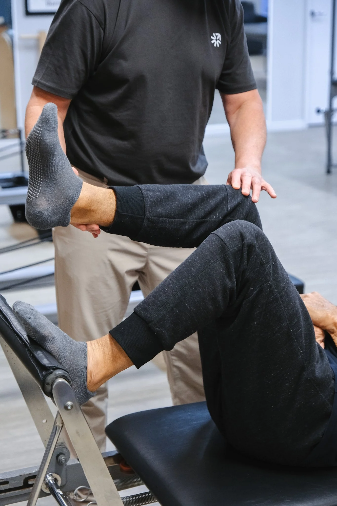 Bunbury Physio - A person in a black T-shirt assisting another person in a black and gray outfit with stretching or leg therapy while seated in a chair with a metal support structure in an indoor setting.