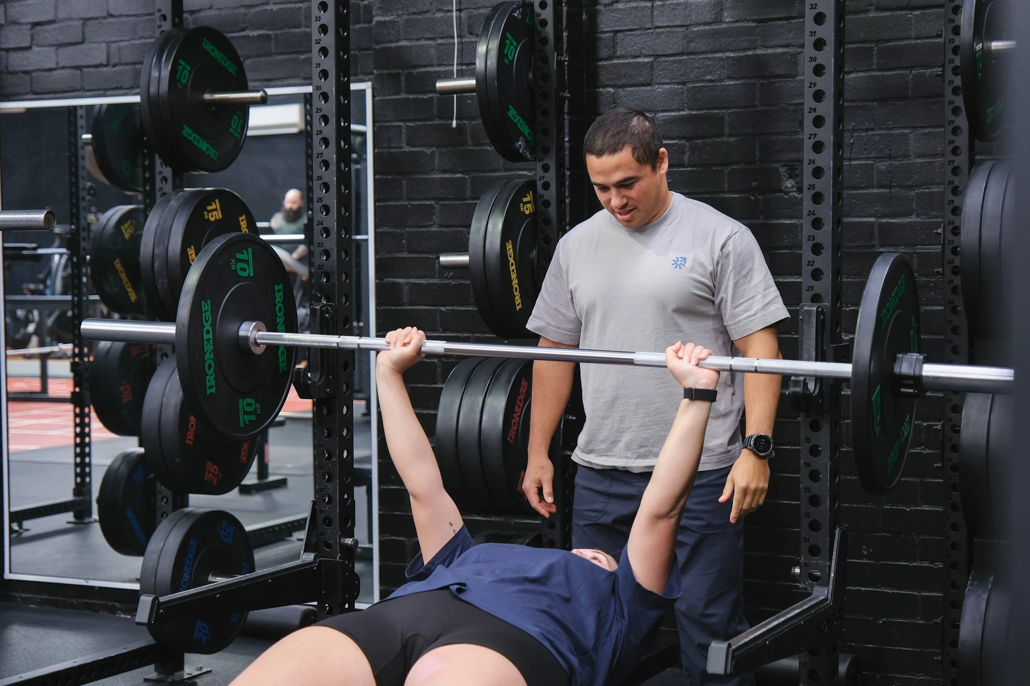 Bunbury Physio - A person lying on a bench press with a barbell, lifting weights with a trainer standing nearby and providing assistance in a gym.