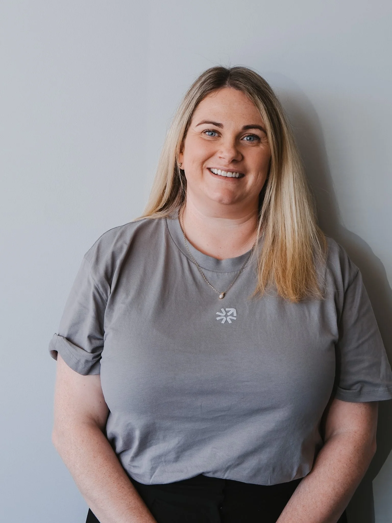 Bunbury Physio - A woman with long hair, wearing a gray T-shirt with a logo, standing against a plain wall, smiling.