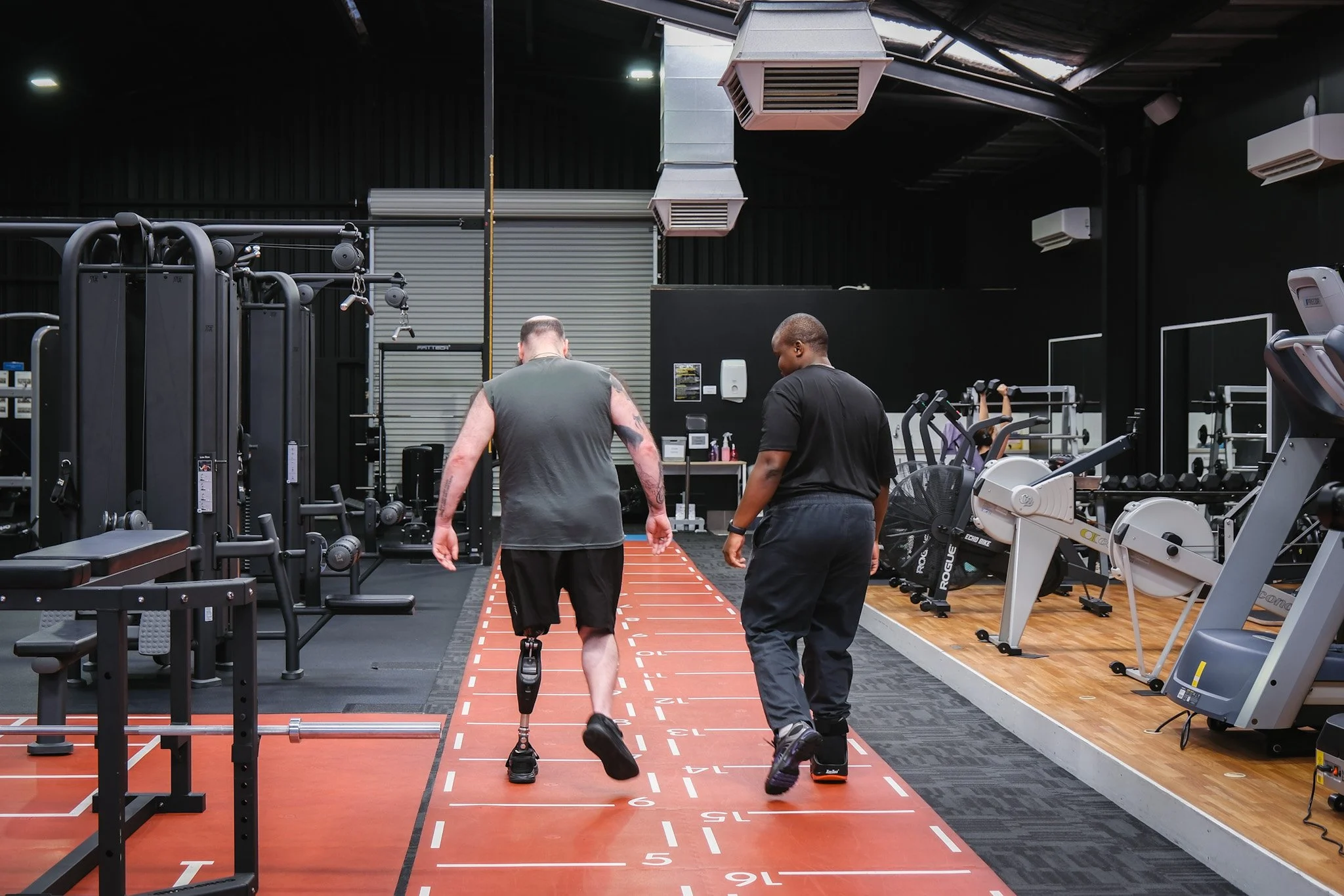 Bunbury Physio - Two men in athletic clothing are walking on a running track inside a gym, with gym equipment and exercise machines visible around them.