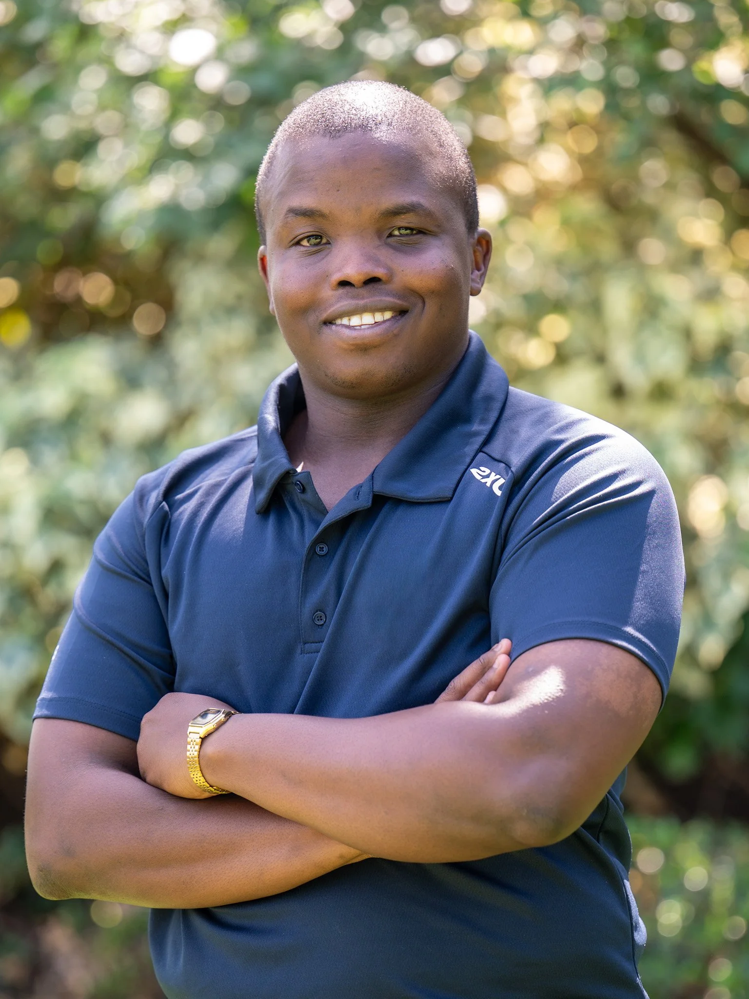 Bunbury Physio - A man with dark skin and short hair standing outdoors with arms crossed, smiling, wearing a navy blue polo shirt with a logo, a gold watch, and a gold bracelet, with a background of blurred green trees and sunlight.