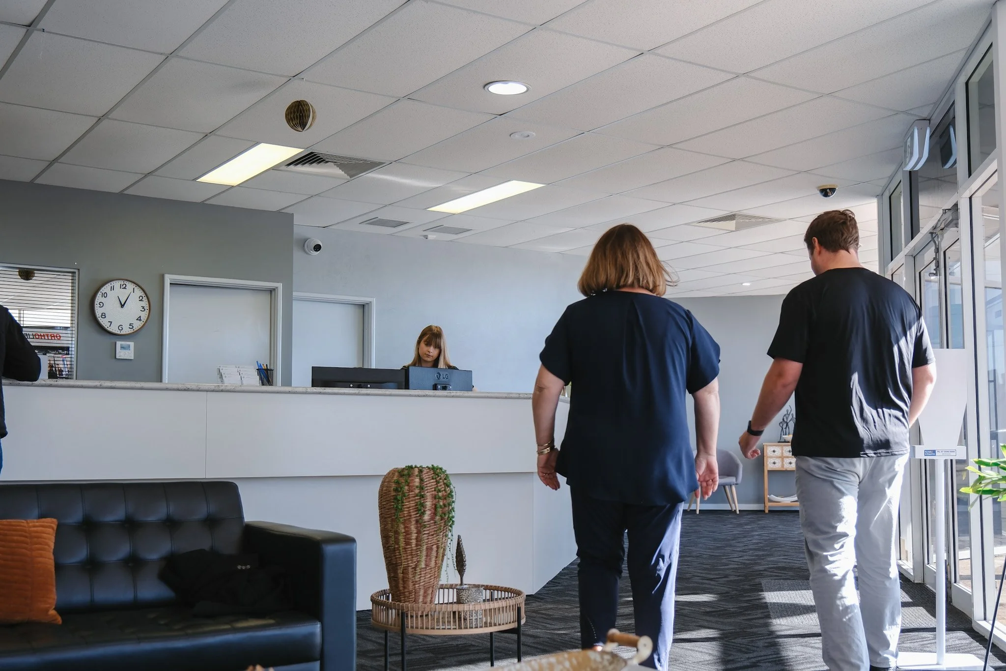 Bunbury Physio - People walking into a modern practice lobby with reception desk, wall clock, and seating area.