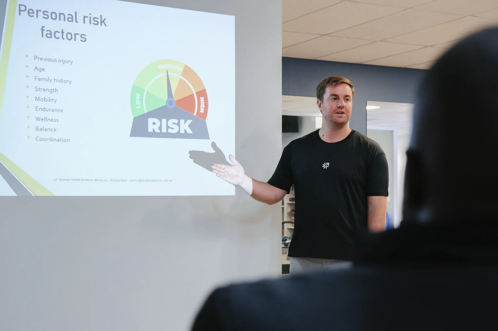 Bunbury Physio - A man in a black shirt presents a PowerPoint slide on personal risk factors in a classroom or conference room setting. The slide includes a gauge chart labeled risk from low to high, with the needle pointing to medium.